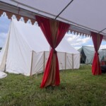 Setup in Courtesan Camp at Pennsic in Æthelmearc on August 30, 2023
<br><br>Photo by Sean O’Fogarty View of a courtyard of a medieval style camp in which are tents and canopies with various furnishings on the ground, and two vehicles
