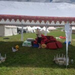 Setup in Courtesan Camp at Pennsic in Æthelmearc on August 30, 2023 View of a courtyard of a medieval style camp in which are tents and canopies with various furnishings on the ground, and a person looking at cushions