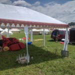 Setup in Courtesan Camp at Pennsic in Æthelmearc on August 30, 2023 View of a courtyard of a medieval style camp in which are tents and canopies with various furnishings on the ground, an SUV, a trailer, and a person looking at cushions