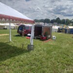 Setup in Courtesan Camp at Pennsic in Æthelmearc on August 30, 2023 View of a courtyard of a medieval style camp in which are tents and canopies with various furnishings on the ground, an SUV, a trailer, and a person walking uphill