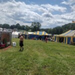 Setup in Courtesan Camp at Pennsic in Æthelmearc on August 30, 2023 View of a courtyard of a medieval style camp in which are tents and canopies with various furnishings on the ground, a trailer, banners on flag poles, and two people walking uphill