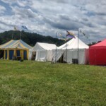 Setup in Courtesan Camp at Pennsic in Æthelmearc on August 30, 2023 View of a courtyard of a medieval style camp in which are tents and canopies with various furnishings on the ground, and banners on flag poles