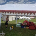 Setup in Courtesan Camp at Pennsic in Æthelmearc on August 30, 2023 View of a courtyard of a medieval style camp in which are tents and canopies with various furnishings on the ground, a person sitting on a pile of cushions, and another person walking under a canopy