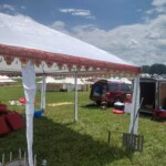 Setup in Courtesan Camp at Pennsic in Æthelmearc on August 30, 2023 View of a courtyard of a medieval style camp in which are tents and canopies with various furnishings on the ground, an SUV, and a trailer