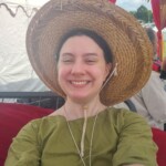 Courtesan Ginevra Fiammetta di Silvestri resting while setting up Courtesan Camp at Pennsic in Æthelmearc on August 31, 2023 Selfie of a woman grinning in a straw hat and green tunic under a fancy canopy in a medieval style encampment