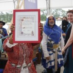Courtesan Ginevra Fiammetta di Silvestri receives her scroll for the Order of the Pearl at Ruby Joust in Atlantia on May 27th, 2023
<br><br>Photo by Llwyd Aldrydd A woman in ancient Roman clothing wearing a red veil smiles and looks to the side while a woman in medieval clothing holds up a medieval style scroll to the camera and others in medieval clothing look on