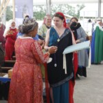 Courtesan Ginevra Fiammetta di Silvestri receives her medallion for the Order of the Pearl at Ruby Joust in Atlantia on May 27th, 2023
<br><br>Photo by Llwyd Aldrydd A woman in ancient Roman clothing wearing a red veil and a gold stag pin being handed a medallion by a woman in medieval clothing wearing a crown while other people in medieval clothing look on