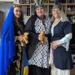 Courtesans Talitha de Barde and Khalida al-Khalilah with Tullian Danyel de Licatia at the Company of the Golden Fox's salon in Caid on February 25, 2023.
<br><br>
Visit the Company of the Golden Fox (Calafia Courtesans Guild) Facebook group here: <a class='blue-link' href="https://www.facebook.com/groups/540612268076159">Facebook link</a> Three people in medieval and Renaissance clothing in a modern living room standing together and smiling at the camera