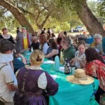 Courtesans Giovanna Ricci and Eleonora di Gerardo teaching Courtesans 101 as part of Courtesan Charm School at Potrero War in Caid on May 26th, 2023.
<br><br>
Visit the Company of the Golden Fox (Calafia Courtesans Guild) Facebook group here: <a class='blue-link' href="https://www.facebook.com/groups/540612268076159">Facebook link</a> Several people in ancient, medieval, and Renaissance clothing gathered around a table outdoors, at the head of which two of them are standing and teaching